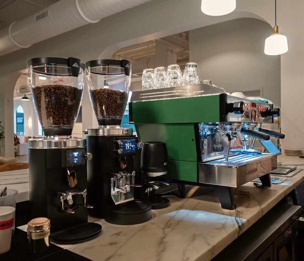 A countertop coffee station setup in a cafe interior, featuring a green and stainless steel espresso machine, two black coffee grinders filled with coffee beans, several clear glass cups, and hanging pendant lights above a marble counter.